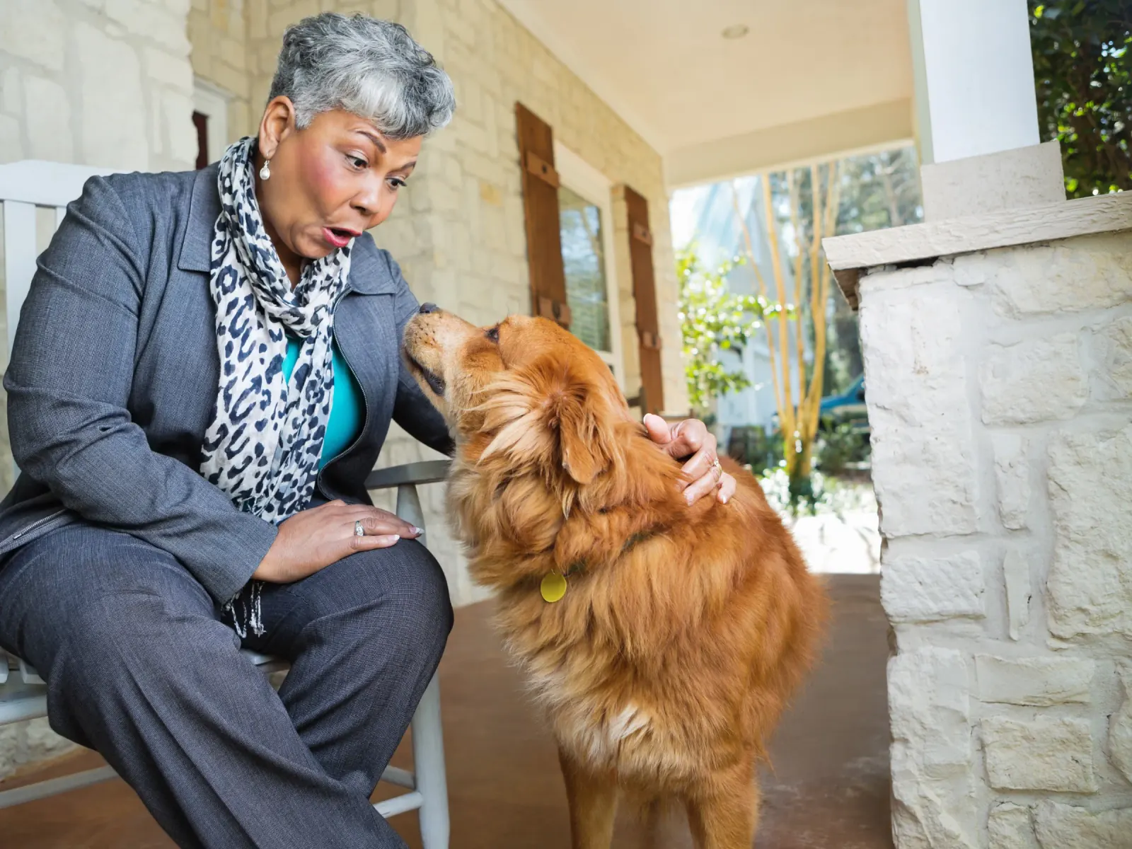 An older woman on her front porch petting a fluffy golden dog